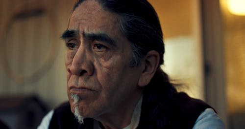 A person with medium skin and long grey-black hair stares to the left of the camera.