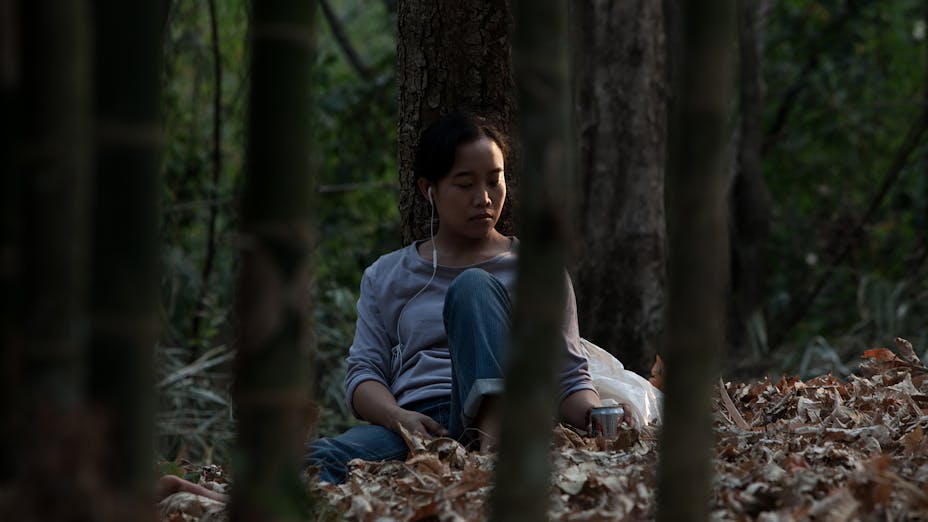 a woman sits against a tree in a wooded landscape listening to music through earphones.