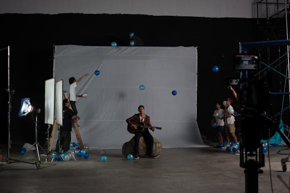 a seated man plays an acoustic guitar in front of a white studio sweep as others around him toss blue balloons.