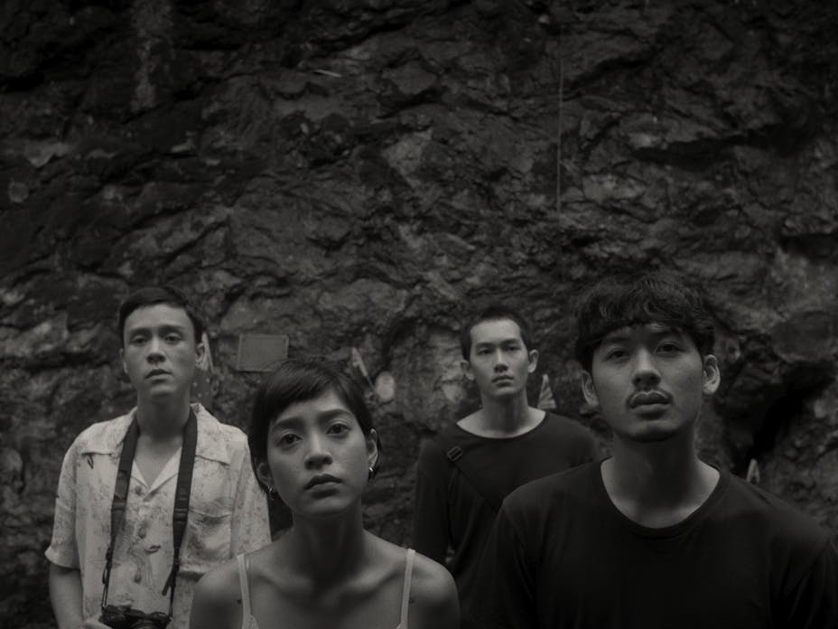black and white image of a woman and three men facing forward in front of a rock wall.