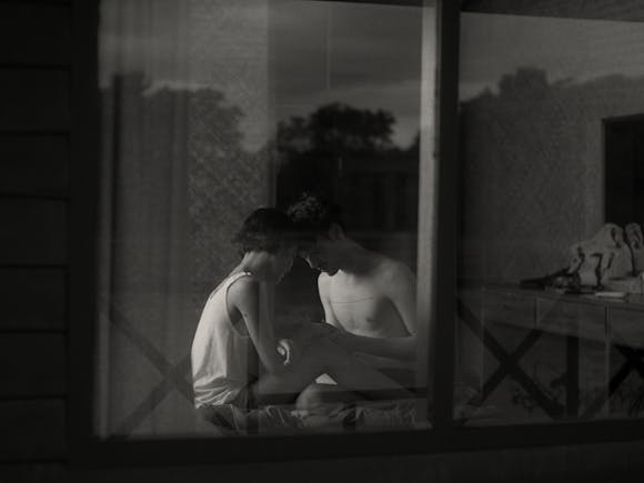 black and white image looking through a window at a couple sitting together in bed embracing.