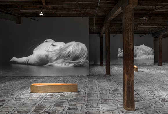 Gallery with white sand on floor with characters drawn in it, small coffins, and video projection of woman on floor