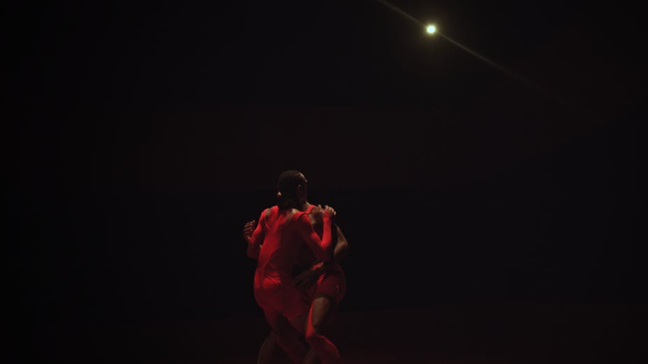 Two dancers with medium-dark skin reach to embrace each other.