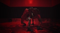 Two dancers with medium-dark skin lunge in opposite directions in a red environment.