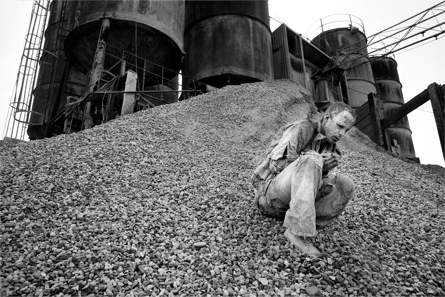 A adult covered in white clay kneels on a massive pile of rocks outside a rusted industrial building.