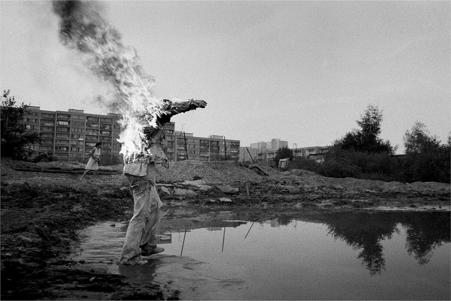 A man on fire walks into a pond outside of a large housing block while a woman in a dress watches nearby.