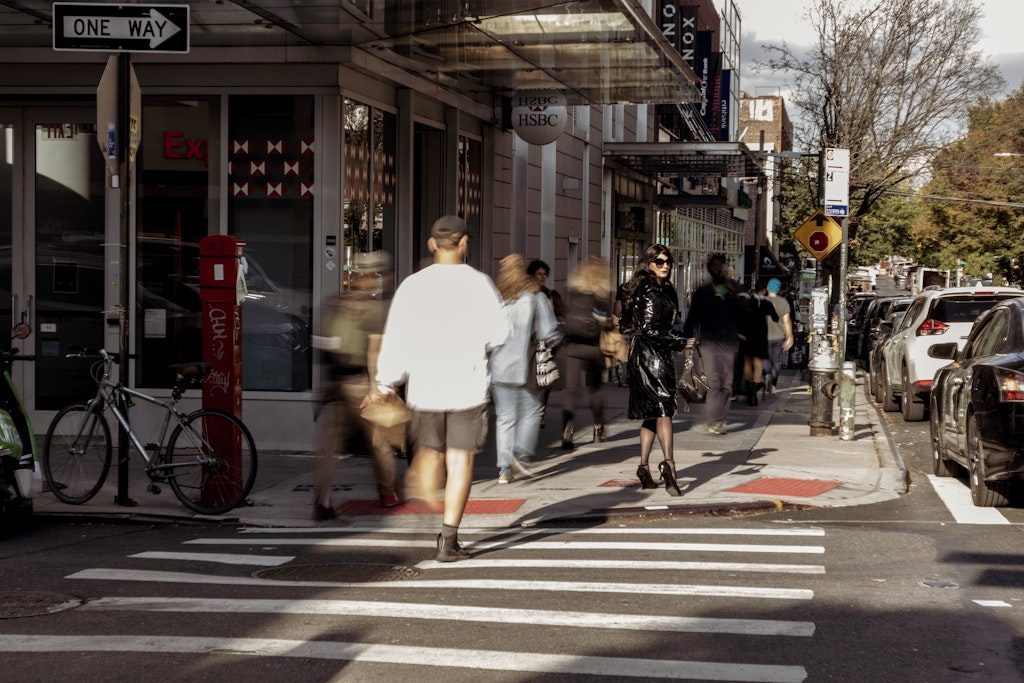 An adult in a black shiny coat and heals is turning to look at the viewer on a busy city street.