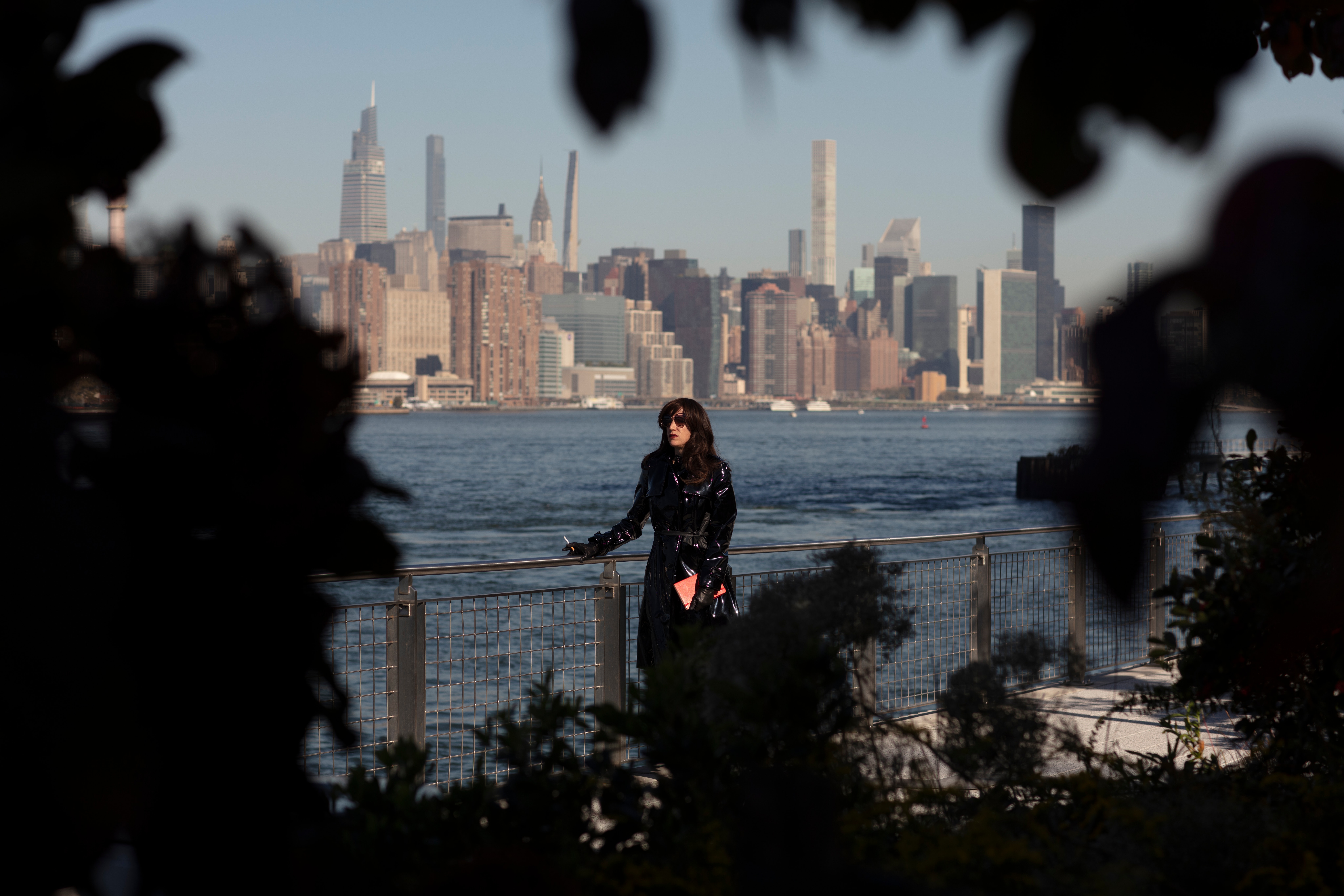 A performer wearing a shiny black jacket and dark sunglasses leans against a waterfront railing. She holds a cigarette in one hand and a red notebook in the other; behind her is the New York City skyline. The image is framed by tree branches and brush, as if taken by someone hiding and observing her from afar.