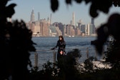 A performer wearing a shiny black jacket and dark sunglasses leans against a waterfront railing. She holds a cigarette in one hand and a red notebook in the other; behind her is the New York City skyline. The image is framed by tree branches and brush, as if taken by someone hiding and observing her from afar.