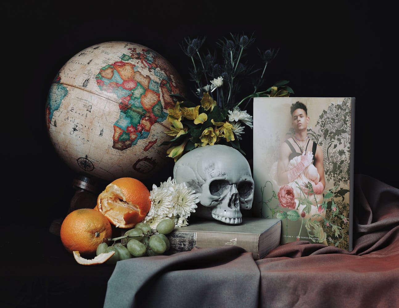 Painting-like still life photograph including skull, globe, oranges, flowers, and magazine cover
