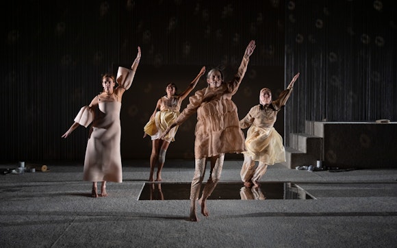 Four performers, each wearing a different, tan, intricately layered dress, dance in unison with their left arms raised, on a carpeted stage.