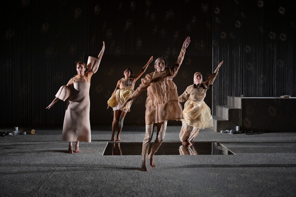 Four performers, each wearing a different, tan, intricately layered dress, dance in unison with their left arms raised, on a carpeted stage.