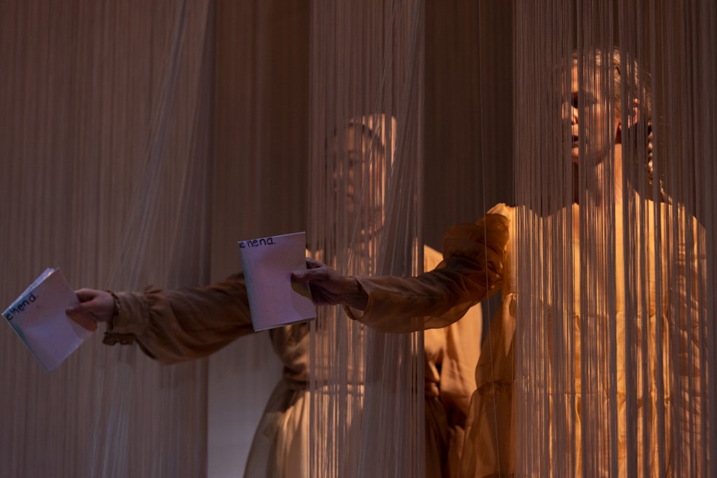 Two performers wearing tan dresses stand obscured behind a gauzy material. Their right arms are extended and holding white folded paper in front of them.