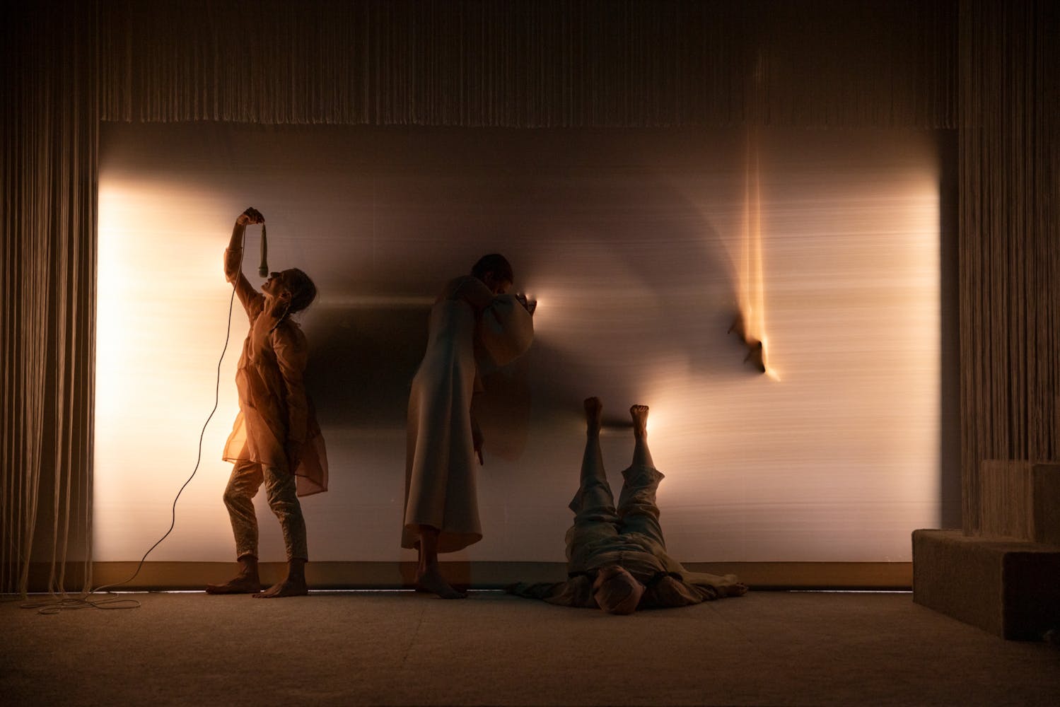 Three figures stand against a blurry wall, one holds a microphone above them, one leans against the wall, the other lays with their legs propped up. All are wearing etheral clothing in shades of light brown.