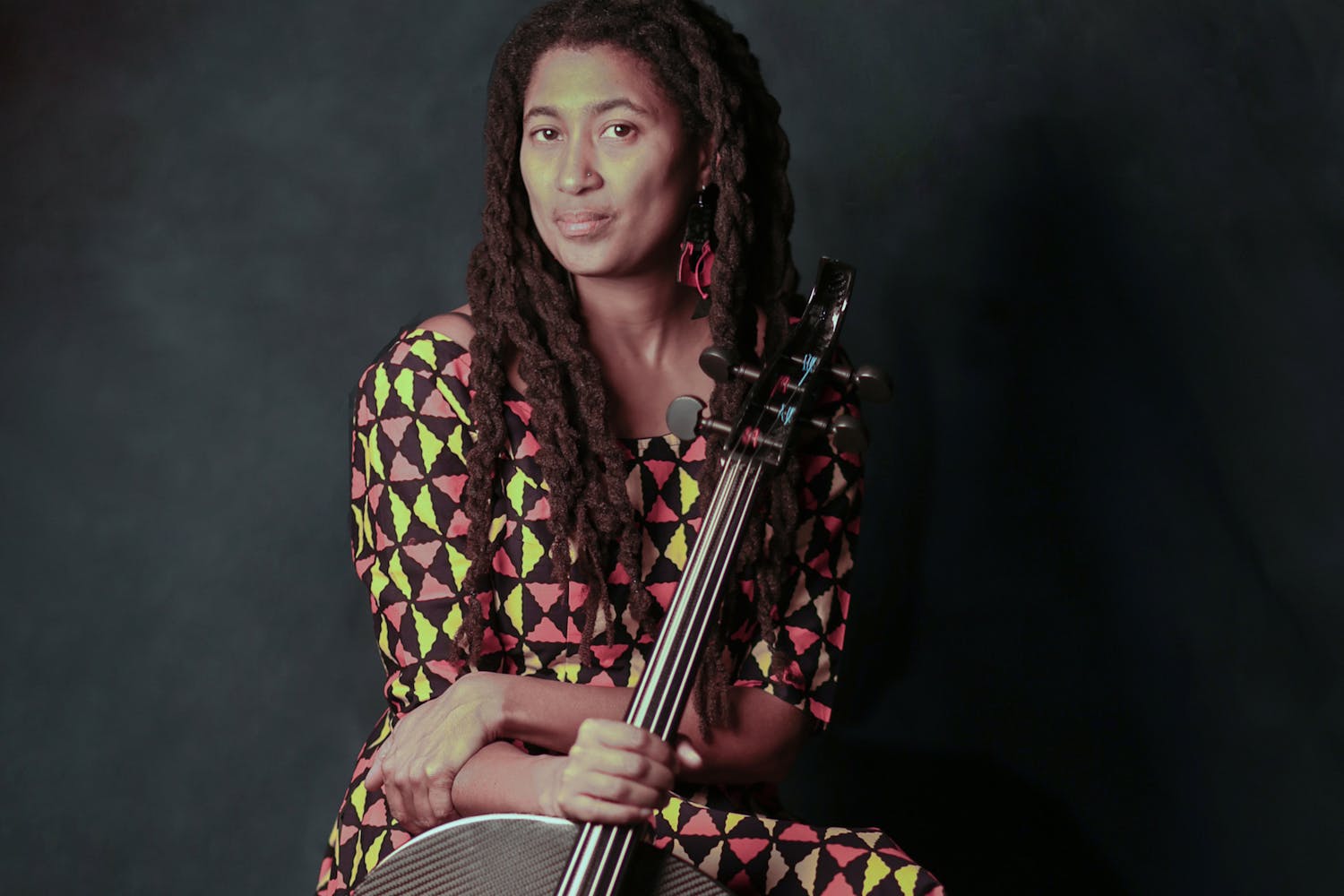 A person with medium-dark skin and long, twisted locks sits wearing a brown dress with yellow and red triangles. They hold their cello with their left hand.