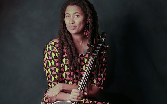 A person with medium-dark skin and long, twisted locks sits wearing a brown dress with yellow and red triangles. They hold their cello with their left hand.