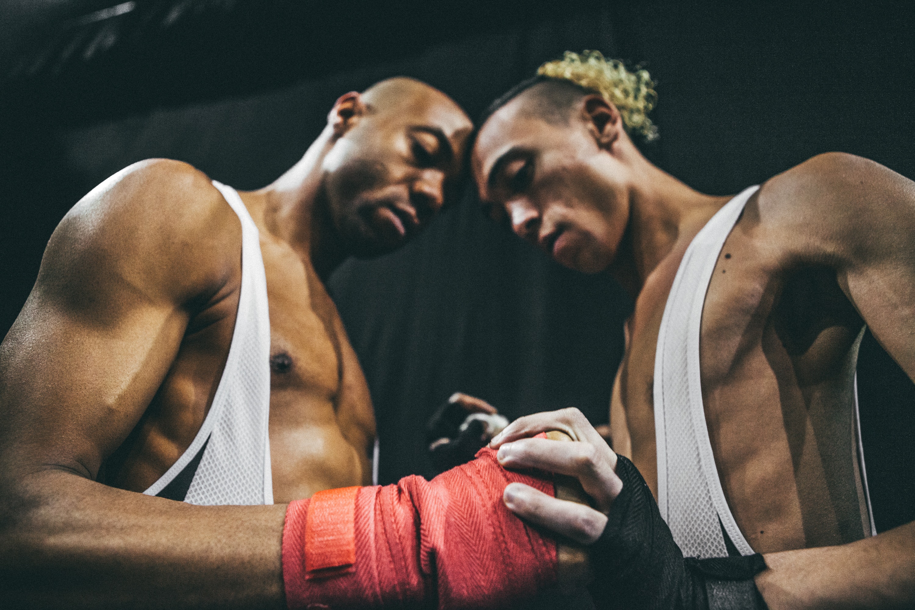 Two adults with dark skin stand with their forheads touching while holding hands, wrapped in boxing tape.