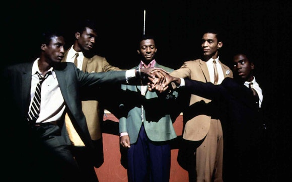 Five men in suits reach out putting their hands together in the center of the group while spotlit on stage.