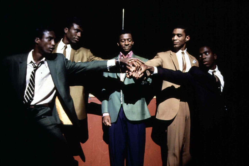 Five men in suits reach out putting their hands together in the center of the group while spotlit on stage.