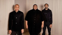 Three musicians dressed in black stand in a diagonal line in front of a wooden background.