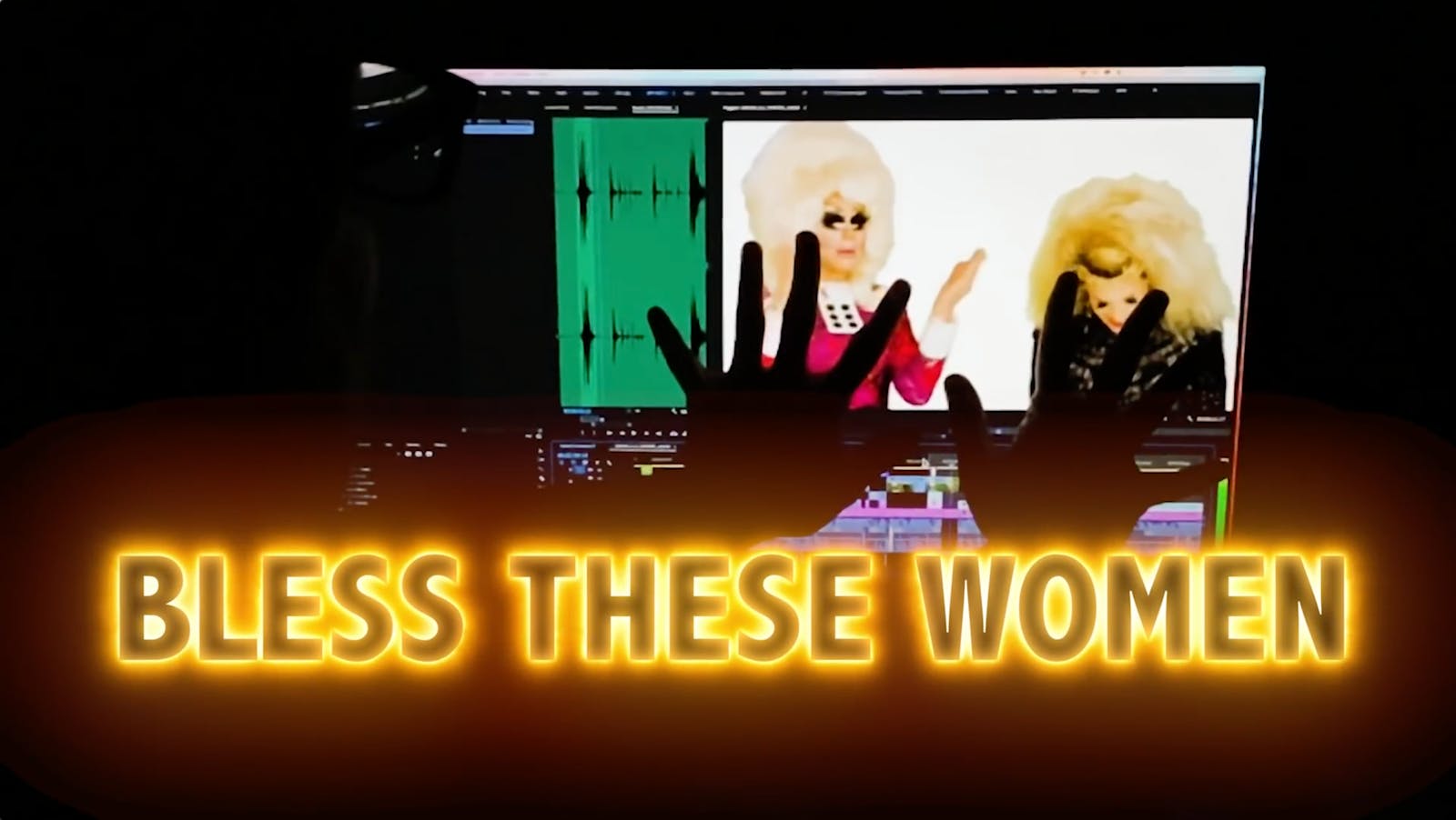 The hands of an editor in a dark room reach up towards a screen with video of two drag queens on it with the text 