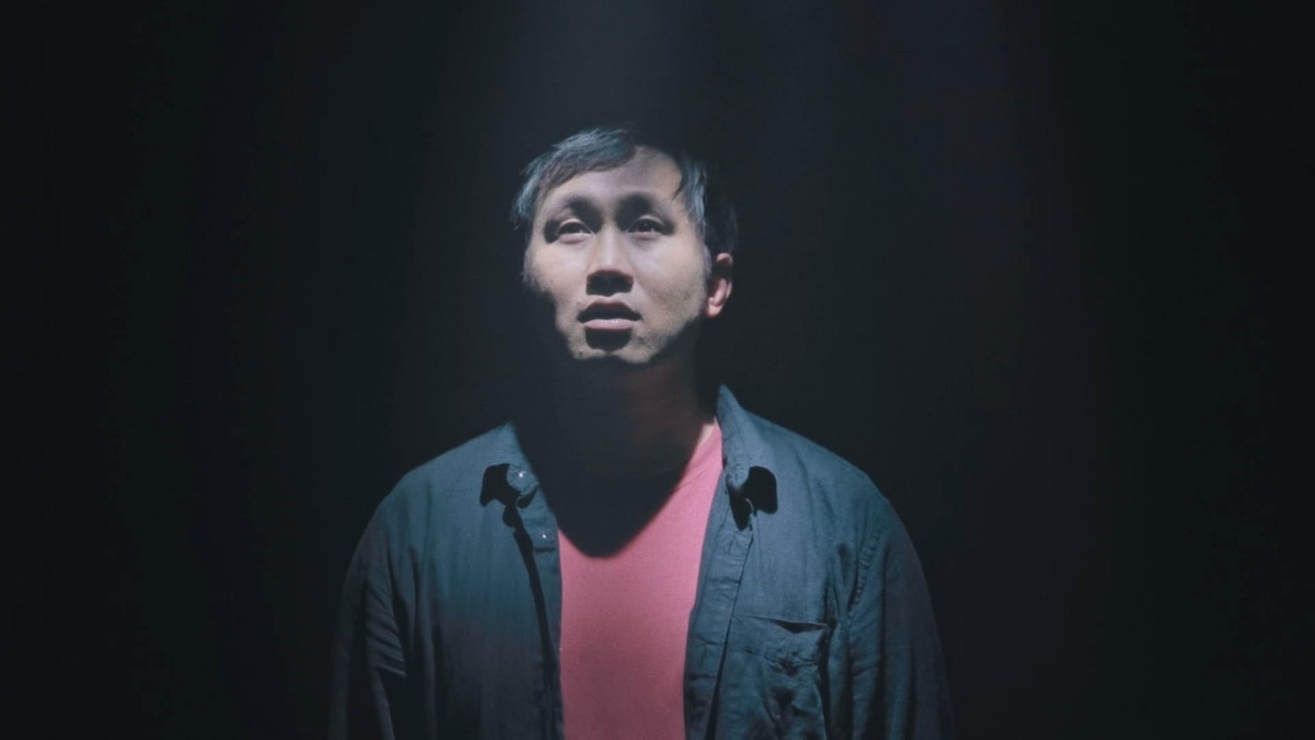 A man is illuminated in the center of a black image. He looks upward with his mouth slightly open.