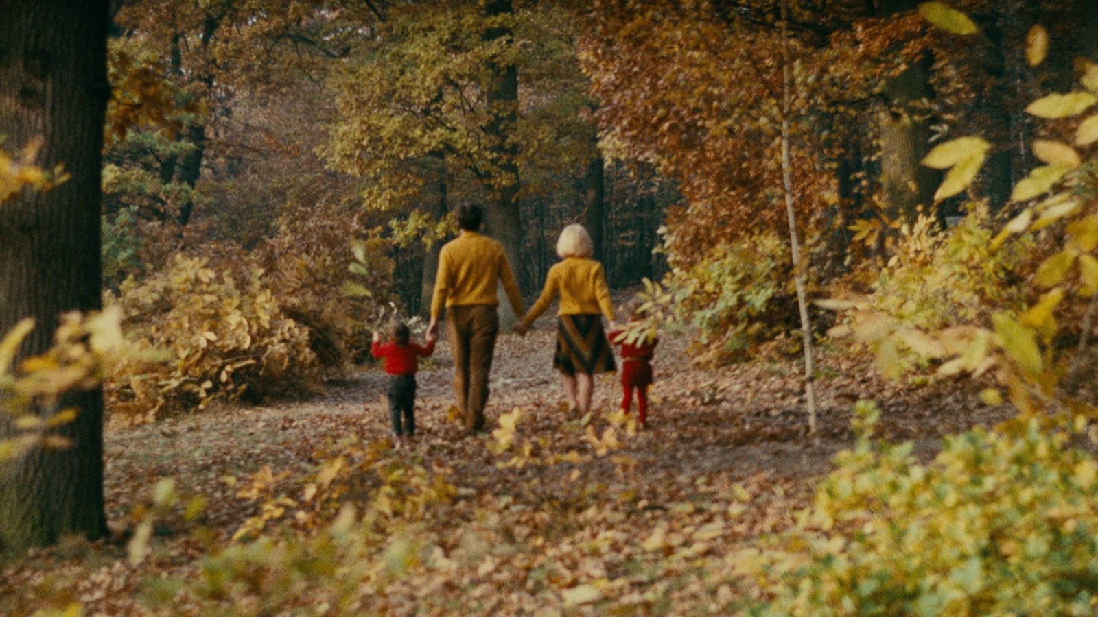 Family of four holding hands and strolling in an autumnal wooded landscape – a man and woman in yellow and two small children in red.