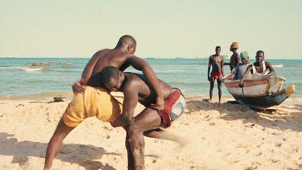 wo men wrestling each other on a sandy beach. A boat on the shore with four others watches on.