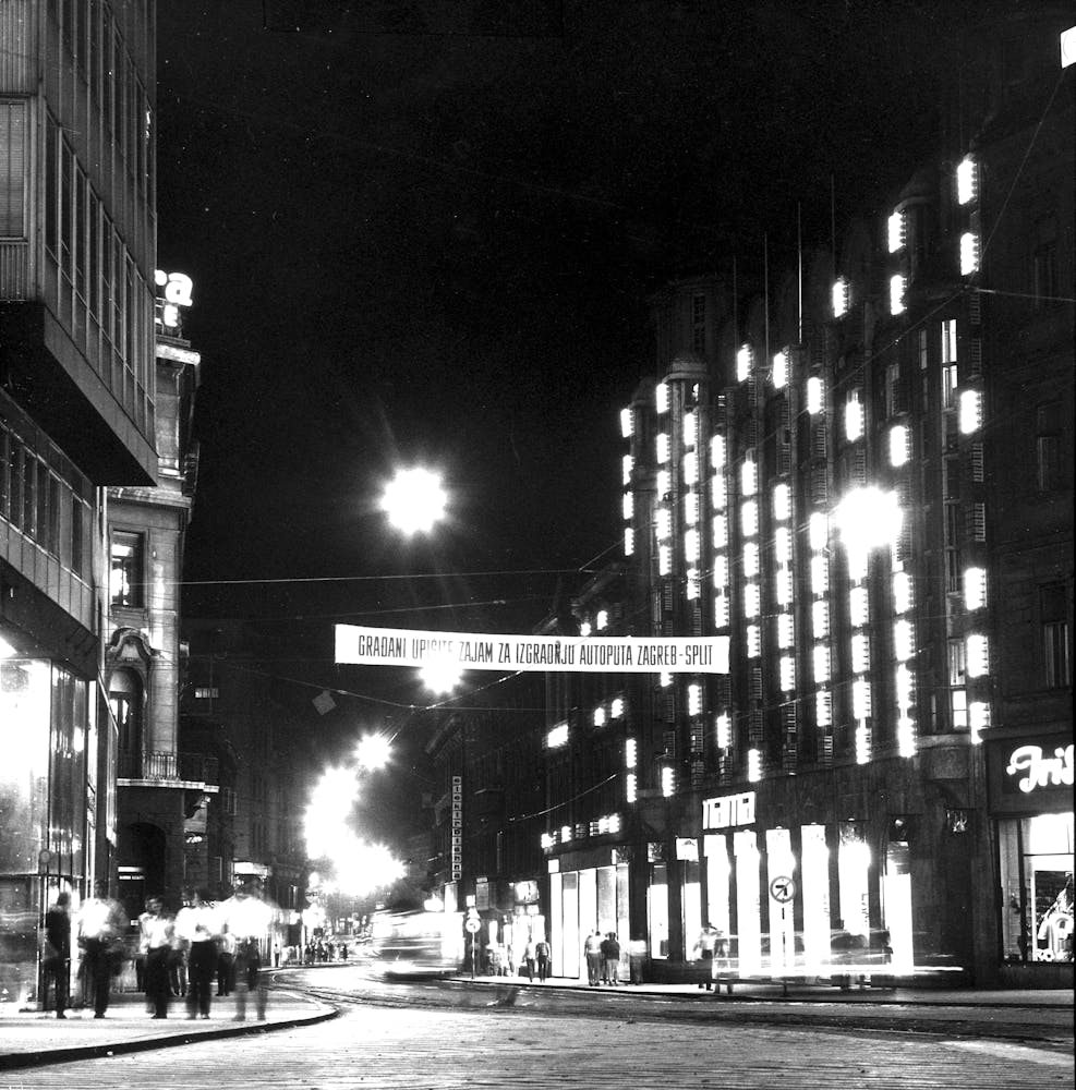 Black and white photograpgh of a building with lights attatched to its exterior at night in a city.