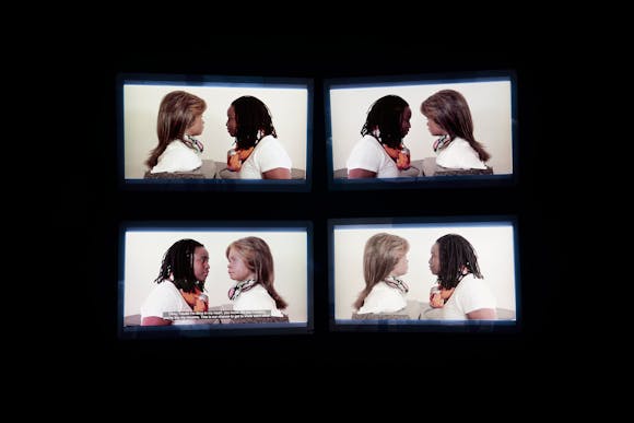 Four monitors on a black wall displaying similar images of a woman speaking face to face with an android head/bust