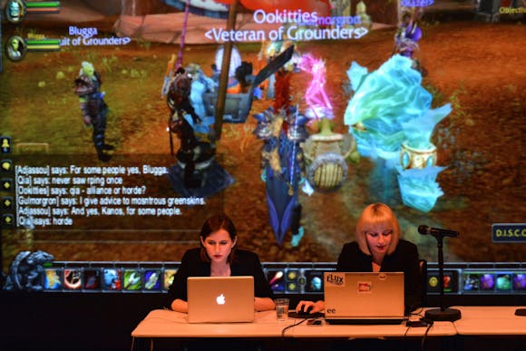 Two women sit at a desk using laptops with a large projection of the video game World of Warcraft behind them.