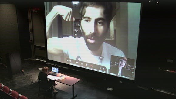 A woman sits at a desk in front of a wall sized projection of a Zoom call with a man flexing his bicep for the camera.