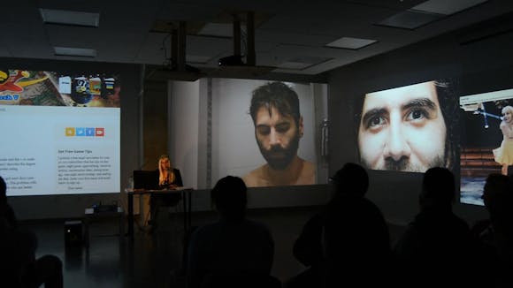 A woman sits at a desk speaking to an audience while using a laptop with projections all around her.