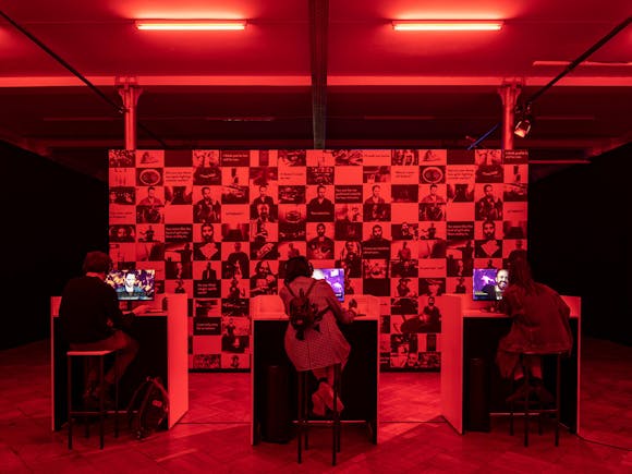 Three adults sit in invidual booths playing a video game in a gallery that is bathed in red light.