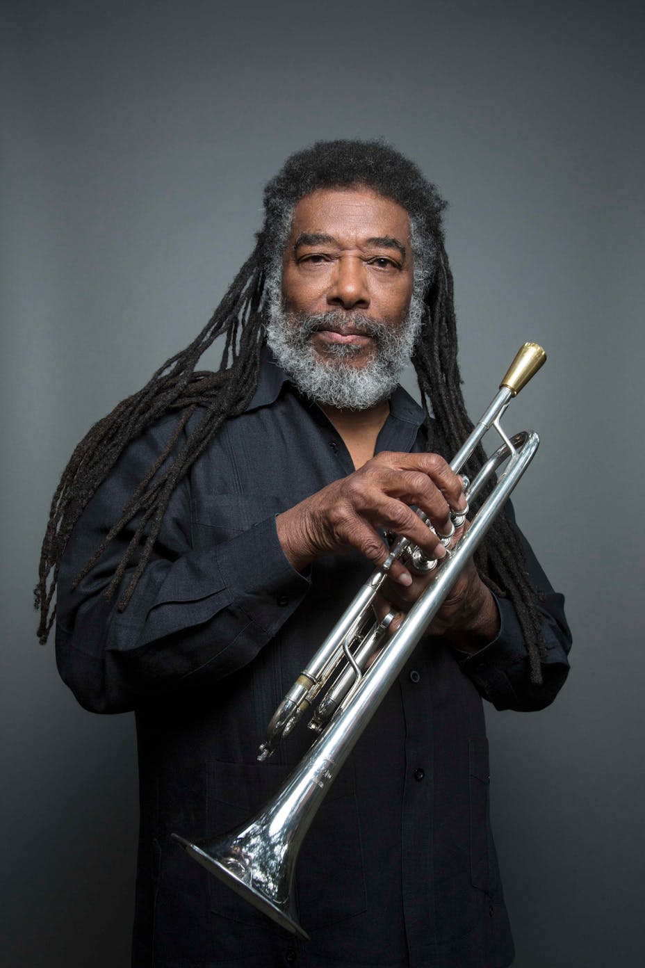 A musician with dark skin and long grey-brown hair holds a silver trumpet.