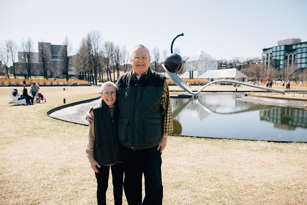 An older couple stand next to one another with their arms around each out in front of a large sculpture of a cherry resting on the tip of a spoon in an outdoor sculpture garden.
