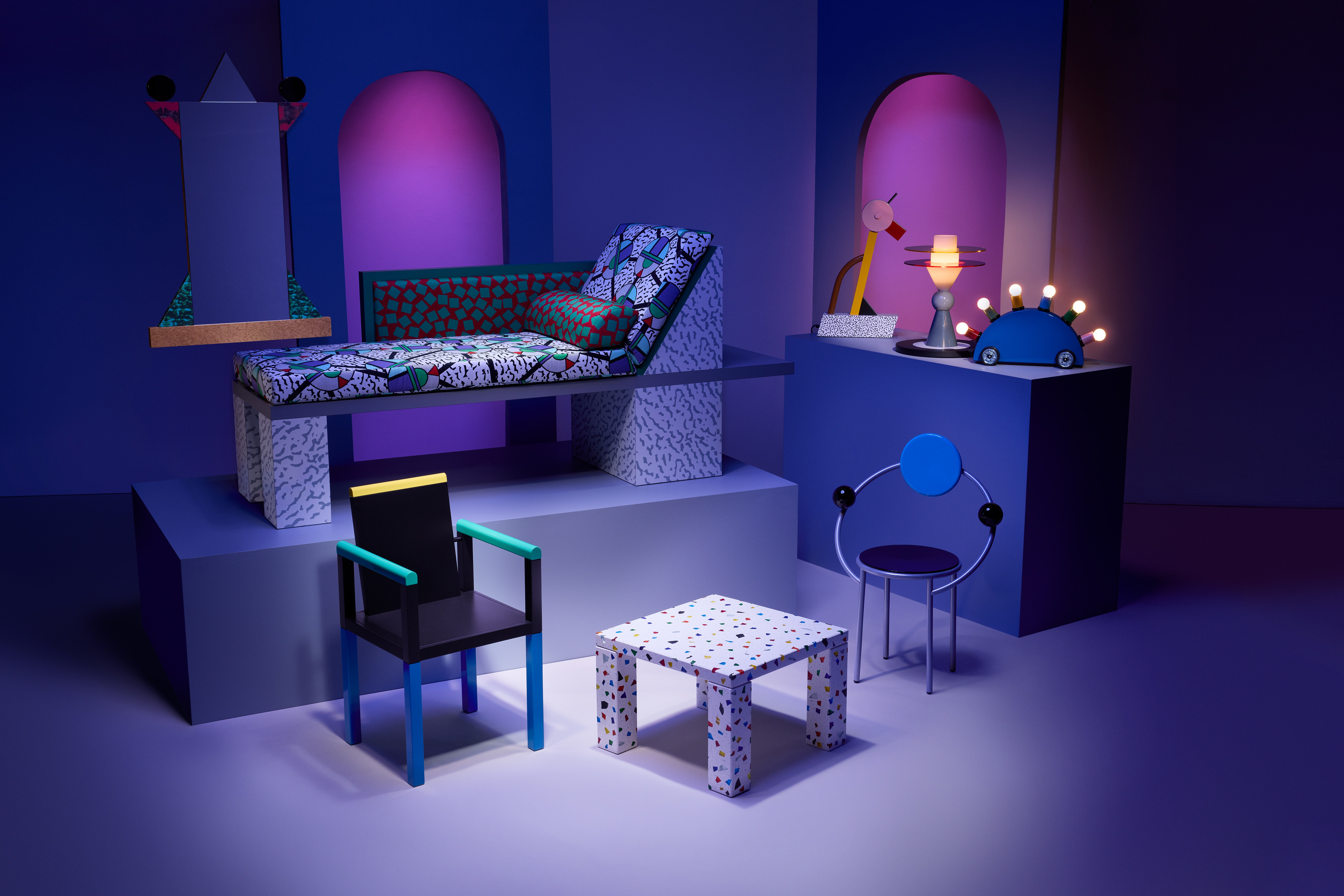 Several pieces of 80's-modern and colorful patterned furniture arranged in a space, lit with blue and purple slights.