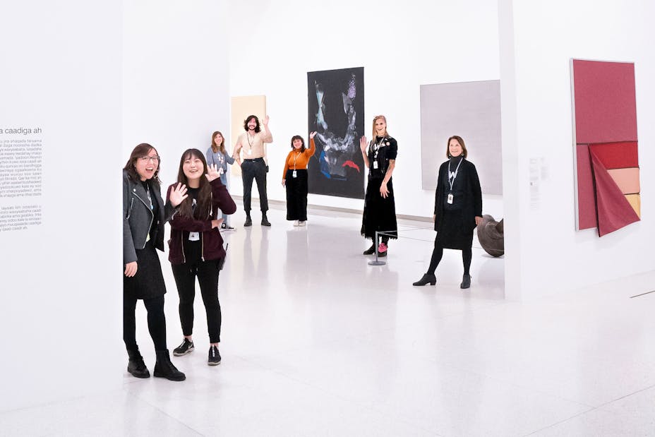 A group of Gallery Assustants wave at the viewer while standing in a gallery of art.