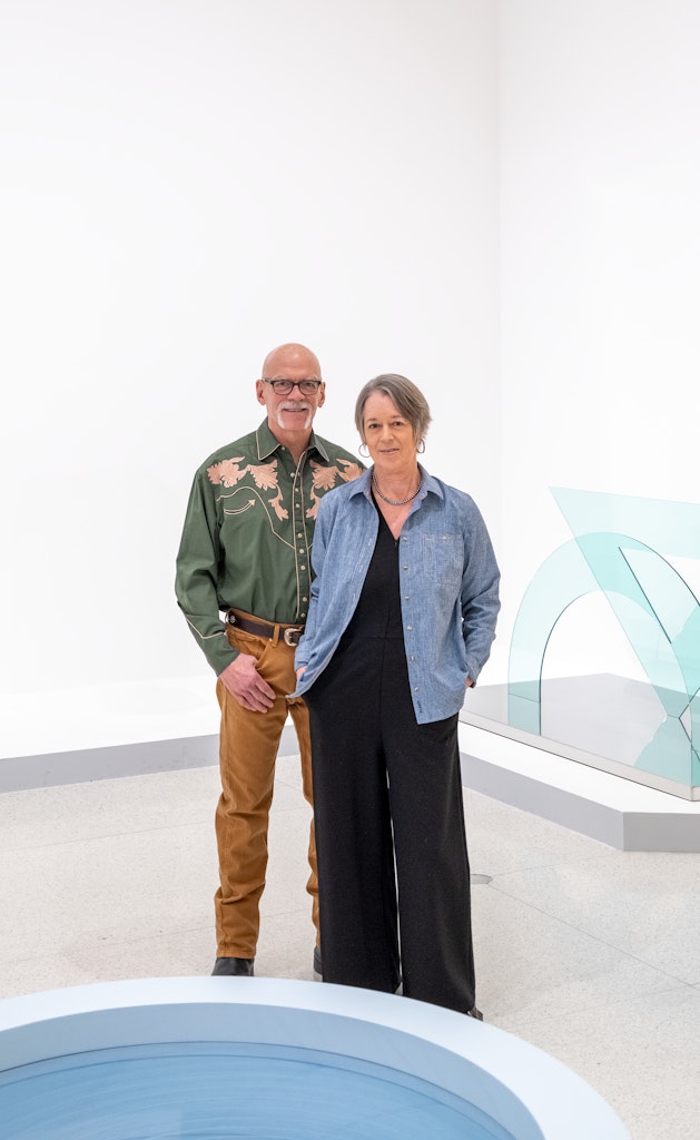 Two adults with light skin, face the viewer smiling, standing in a white gallery space. One has glasses and is bald and one has short grey hair and a denim shirt.