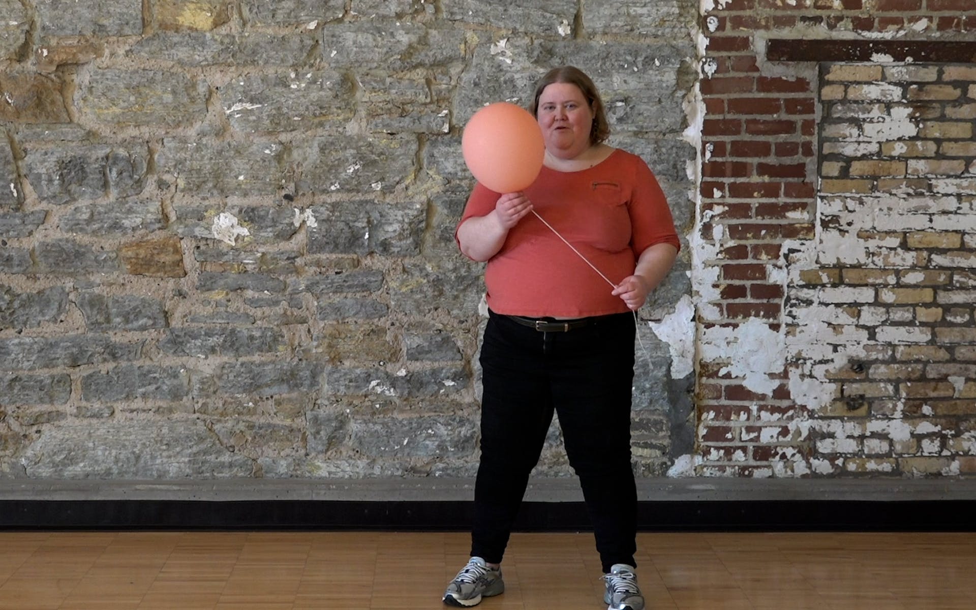 Alison Bergblom Johnson, a plus-size woman with hair just above her shoulders, wearing a casual peach top and jeans with sneakers holding a peach balloon attached to a length of kitchen twine.