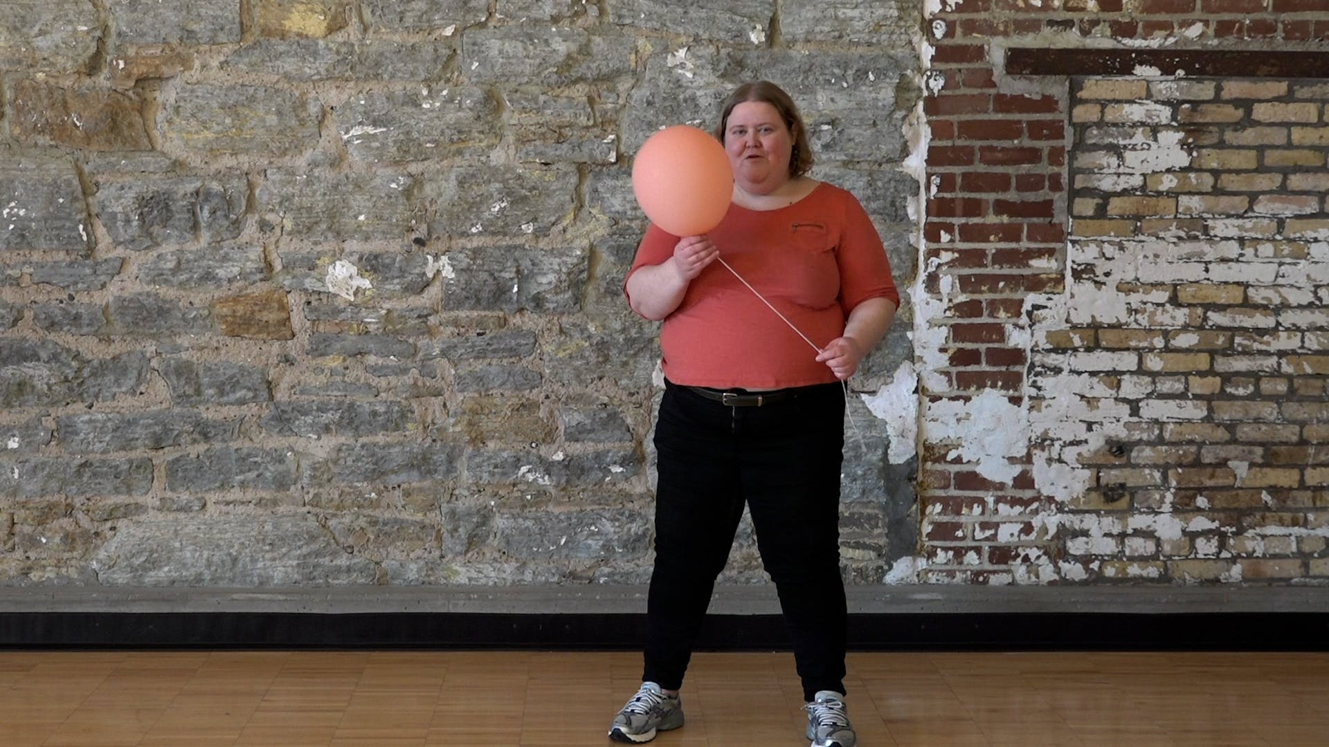 Alison Bergblom Johnson, a plus-size woman with hair just above her shoulders, wearing a casual peach top and jeans with sneakers holding a peach balloon attached to a length of kitchen twine.