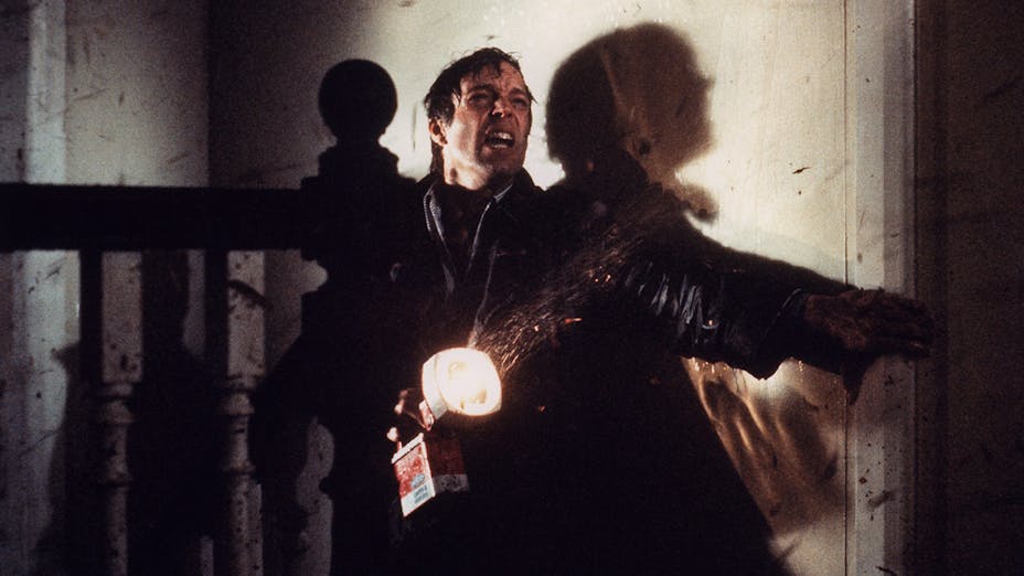 A man with light skin squints with a strained expression and grasps a doorway while holding a flashlight in a rainstorm.