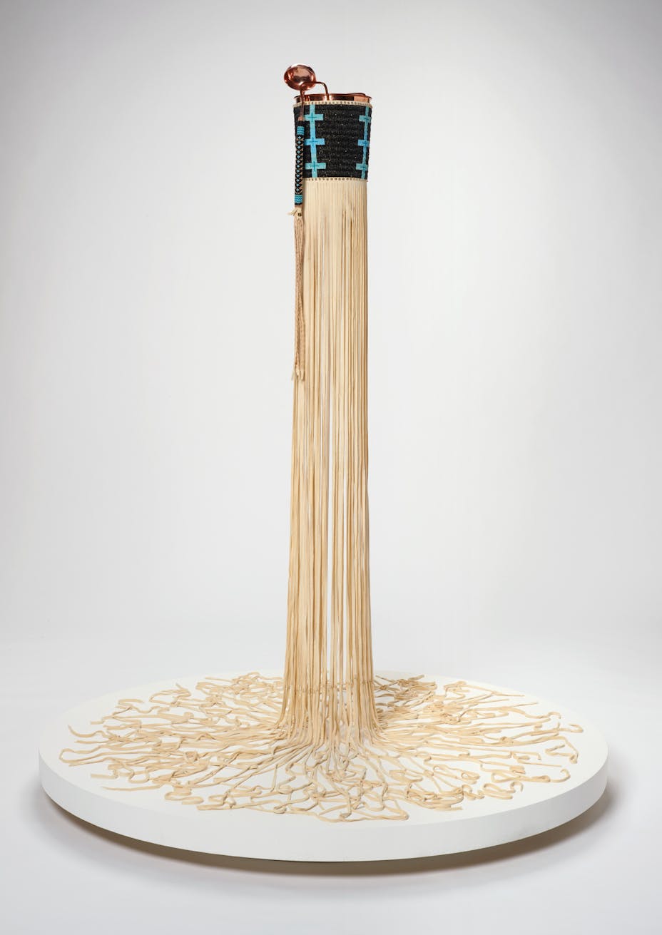 At the upper center of the image a copper bucket and ladle covered in black and blue beadwork are visible, from the bases of the bucket is a cascade of cream colored buckskin fringe which runs vertically onto a white circular platform base. The fringe radiates outwards in squiggling directions from the center.