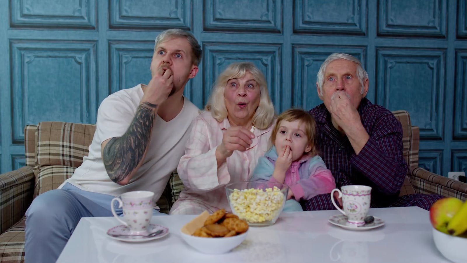 An adult man, a senior man, a senior wonman, and a child eat popcorn together and gaze out in the same direction.
