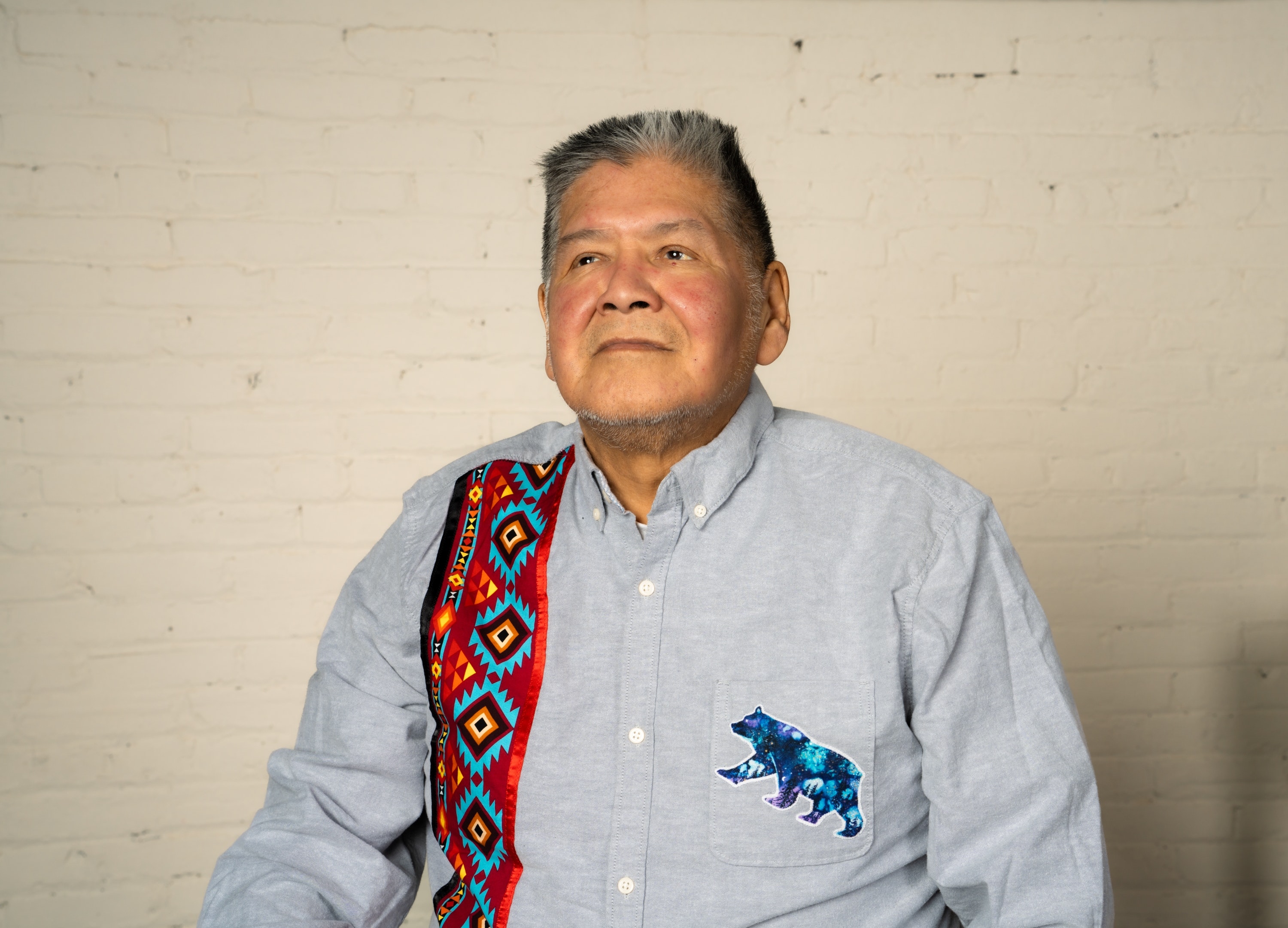 An adult with medium tan skin and short dark hair with gray streaks wears a light blue shirt with southwestern print sits in front of a white bring wall facing the viewer.