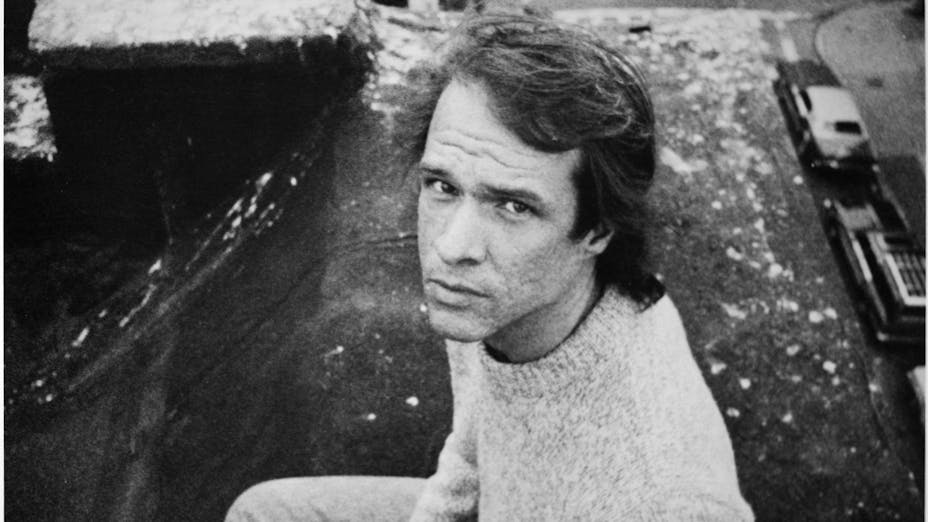 Black and white photo of a man in a sweater looking up at the camera while sitting on a rooftop ledge.