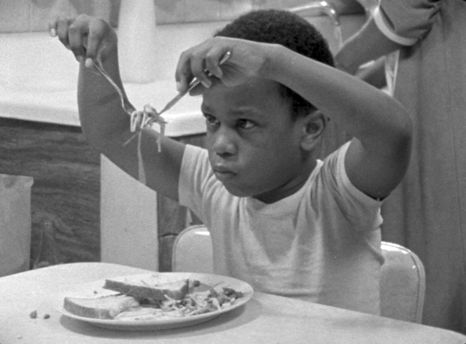 Black and white still of a little boy sitting at a dining table holding up noodles with a fork and knife in front of his face and staring at it.
