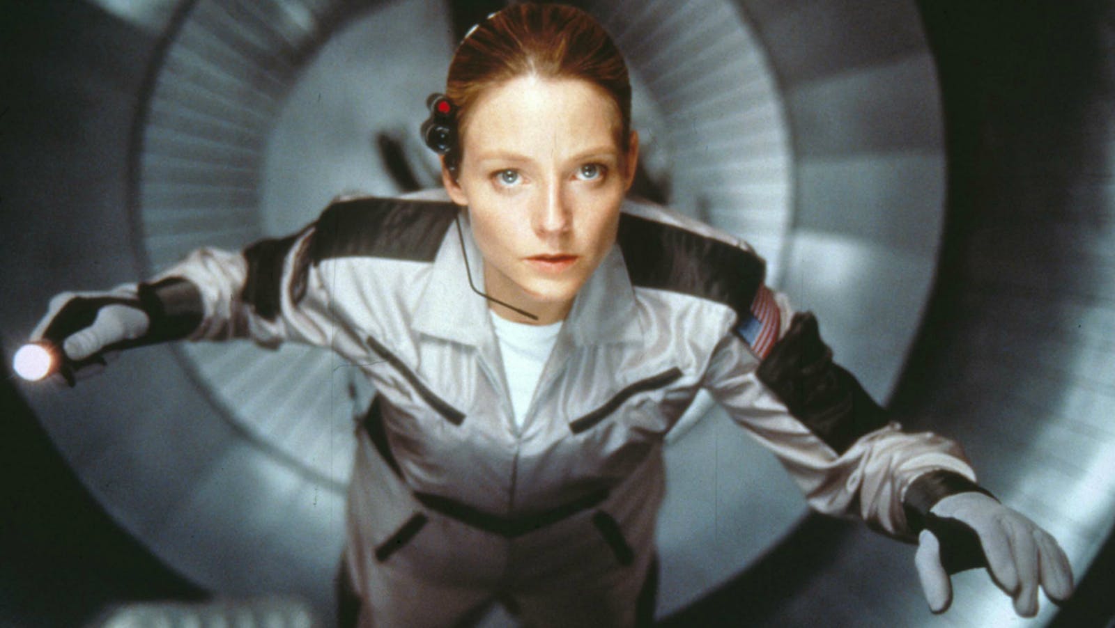 A woman with light skin (Jodie Foster) wearing a space suit with gloves and a headset looking up from within a silver tunnel of a space ship.