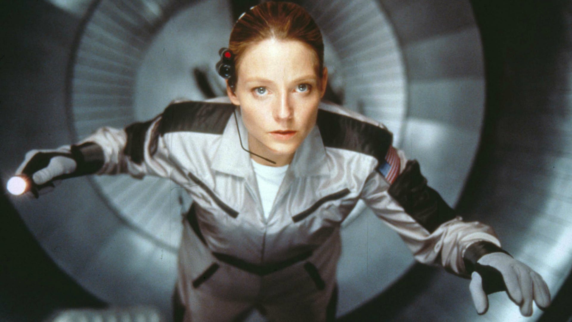 A woman with light skin (Jodie Foster) wearing a space suit with gloves and a headset looking up from within a silver tunnel of a space ship.
