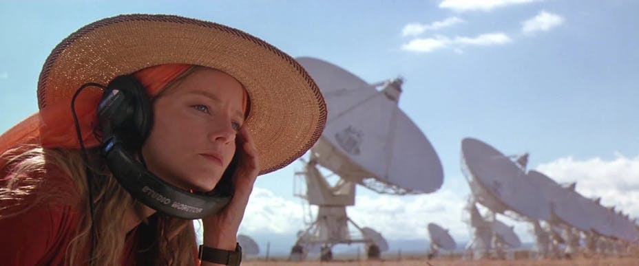 A blonde woman with light skin (Jodie Foster) wearing a straw hat and listening intently to headphones sits in a field of giant satellite dishes.